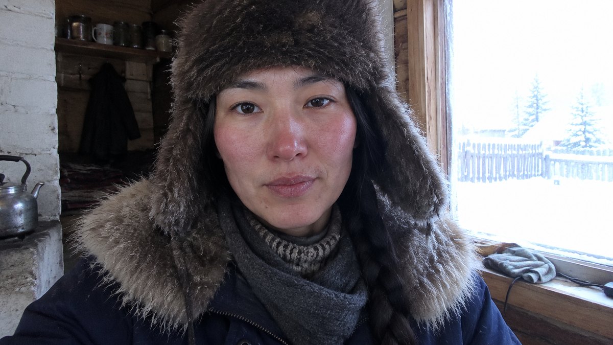 Sardana in a fur hat and parka inside her wooden house, snow in the window behind her